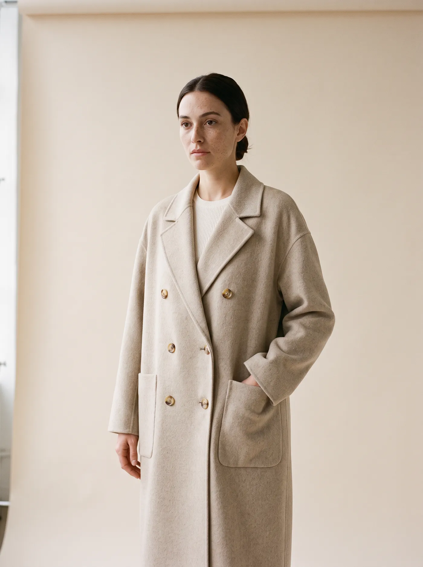 A woman in the long oat Olmo coat, one hand in its patch pocket, standing by a tall window.
