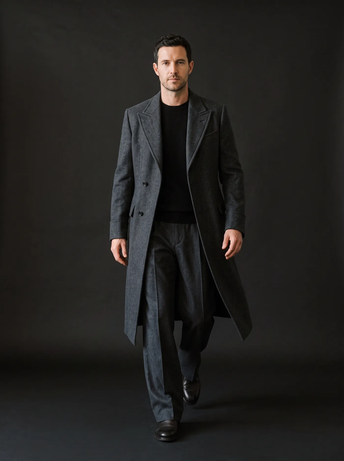 A man walking toward the camera in the charcoal Tejo greatcoat over wide charcoal trousers, against a dark backdrop.