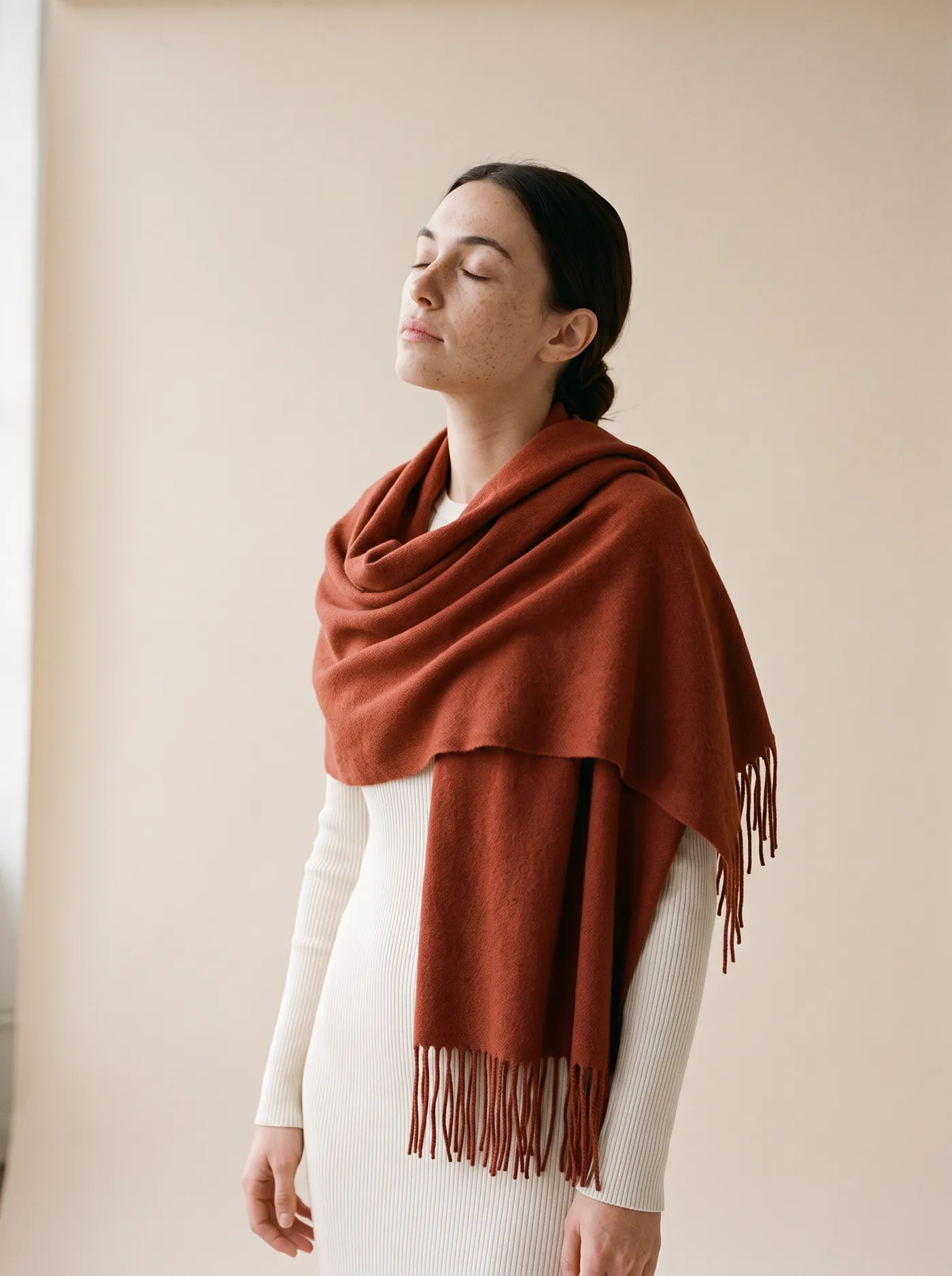 A woman with her eyes closed, a rust cashmere wrap folded around her shoulders over an ivory knit.